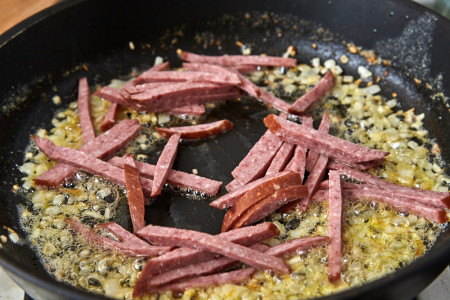 Поки цибуля обсмажується – наріжте 100 гр варено-копченої ковбаси соломкою. Додайте до цибулі, обсмажте помішуючи близько хвилини, а потім зніміть сковороду з вогню.