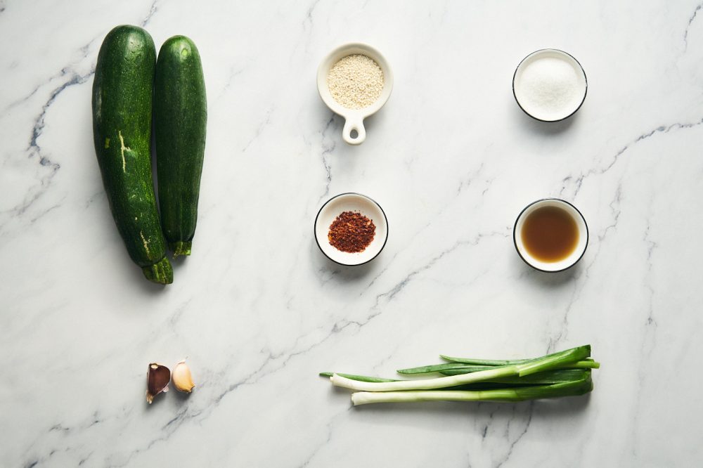 Ingredients for making Korean style zucchini salad: zucchini, garlic cloves, green onion stems, fish sauce, sugar, chili flakes, sesame seeds, sesame oil and vegetable oil