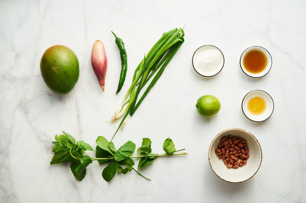 Ингредиенты необходимые для приготовления тайского салата с зеленым манго: манго зеленый, перец чили, лайм, луковица шалота, стебли зеленого лука, рыбный соус, сахар, кунжутное масло, арахис, мята. 